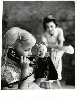 [Blonde girl holding telephone and puppet while mother watches]