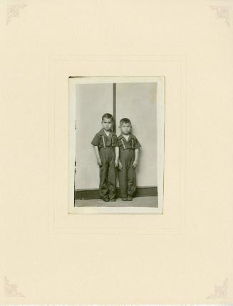 Two standing boys in matching suspenders, striped background