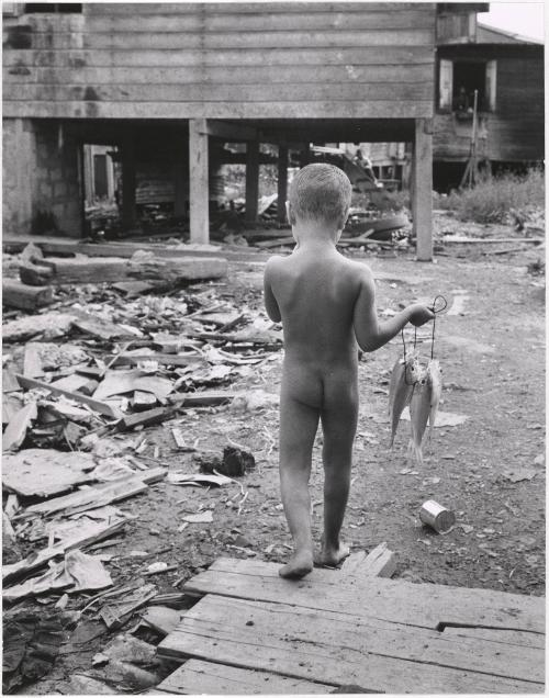 Unclothed boy carrying fish walking through debris, Puerto Rico