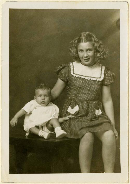 Girl in polka-dot dress with infant sibling supported by metal harness, both seated on table