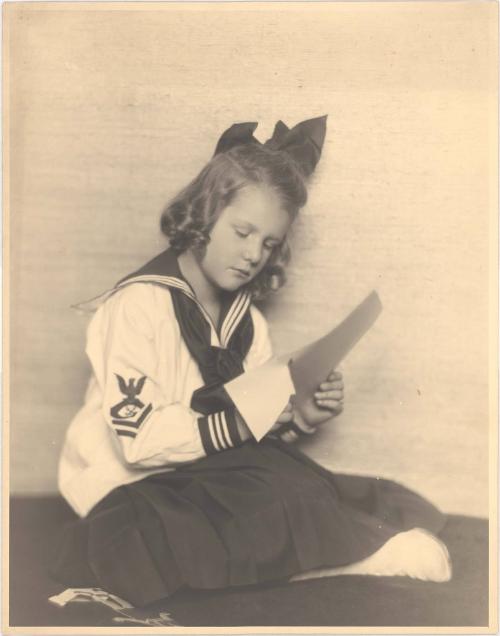 Portrait of seated girl in sailor dress, bow in hair, cutting with scissors