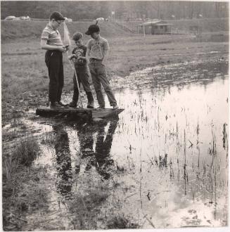 Boy with glasses and two playmates playing on wooden crate in lake from the series Big Brother