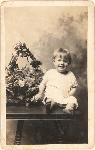 Happy toddler seated on table with basket of flowers, cloud background