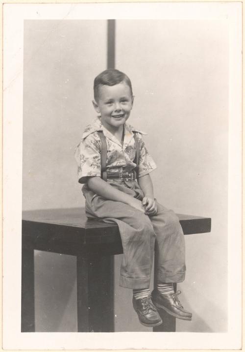 Boy in overalls seated on table, striped background